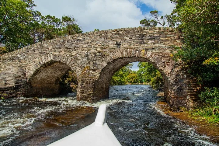 View from the front of a white boat approaching a double-arched stone bridge over rapids