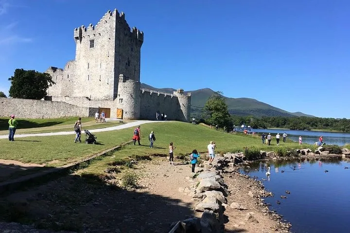 Tourists walking on a grassy path near a stone castle bordering a lake
