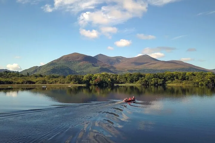 Small boat with passengers traveling across a calm lake reflecting large mountains under blue sky