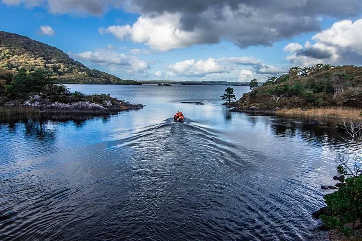Boat moving across calm blue lake water towards distant mountains under a cloudy sky